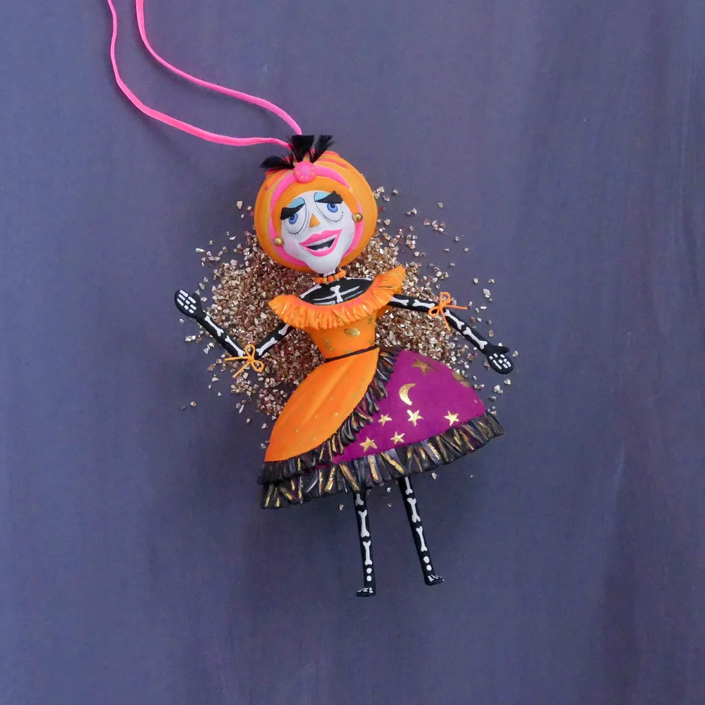 Glitterville Nelly Skelly Fortune Telly hand-painted Halloween ornament featuring a colorful skeleton character in an orange and purple dress, laying on a bed of glitter.