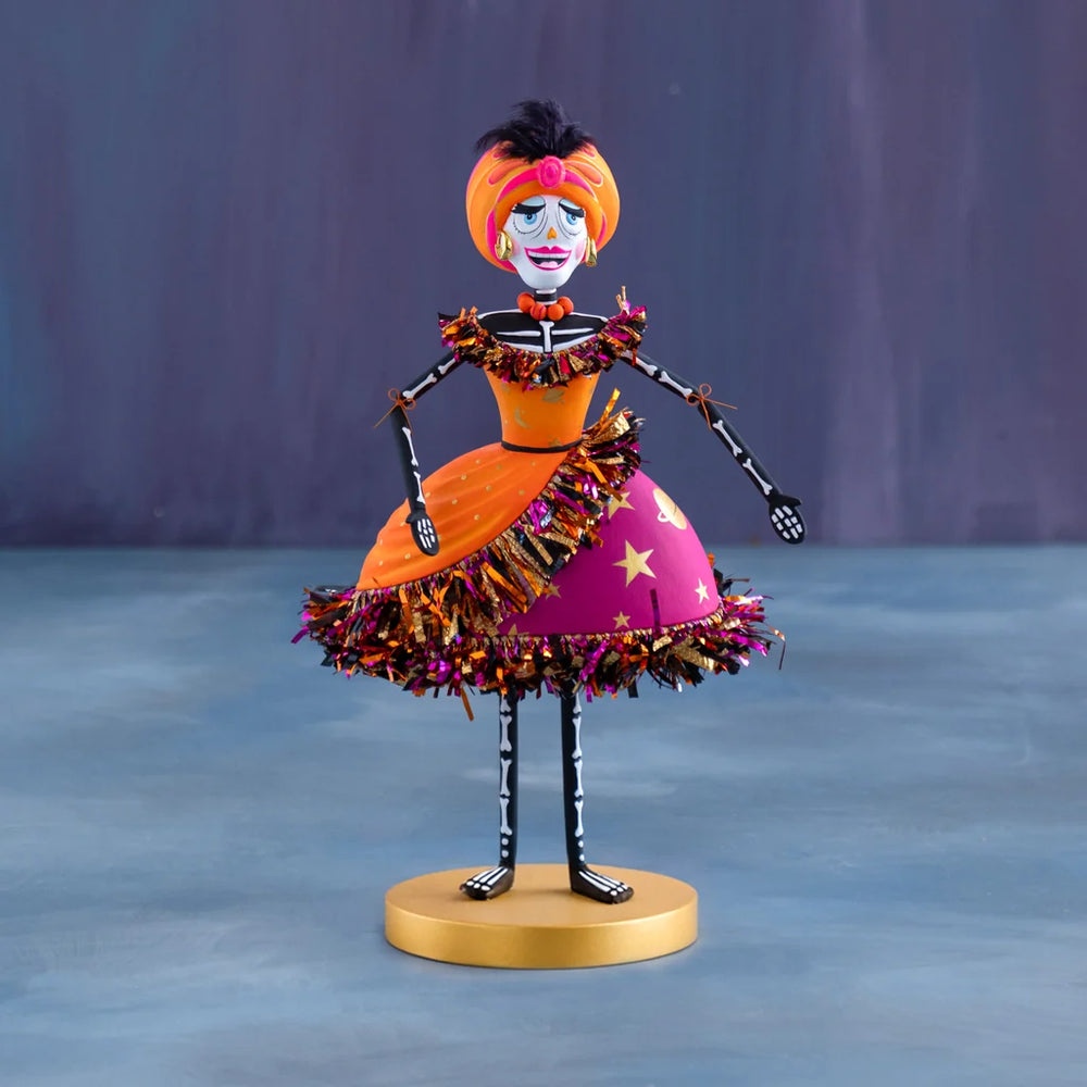 Glitterville Nelly Skelly Fortune Telly tabletop display featuring a hand-painted skeleton figure in a vibrant orange and hot pink dress with metallic fringe trim, standing on a gold round base.