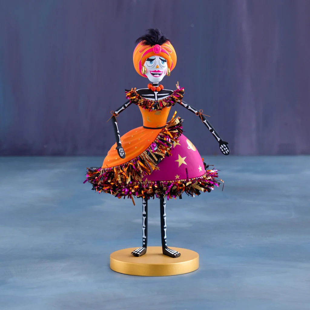 Glitterville Nelly Skelly Fortune Telly tabletop display featuring a hand-painted skeleton figure in a vibrant orange and hot pink dress with metallic fringe trim, standing on a gold round base.