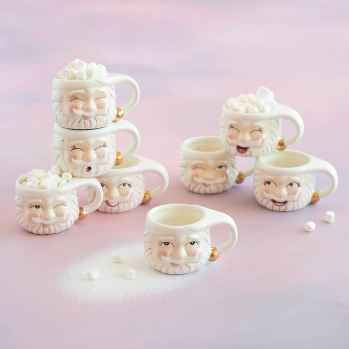 Glitterville Papa Noel ceramic Santa mug set of 8, featuring white Santa face mugs with gold detailing and eight different cheerful facial expressions, with some mugs topped with marshmallows.