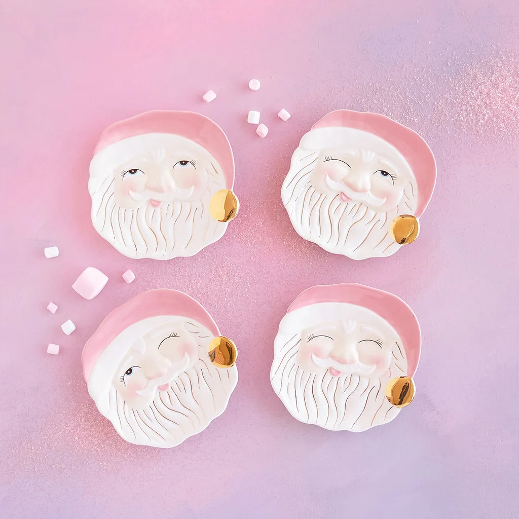 Glitterville Papa Noel pink Santa Claus cookie plate set featuring four ceramic Santa face plates with pink hats, gold bell details, and different facial expressions on a pink background.