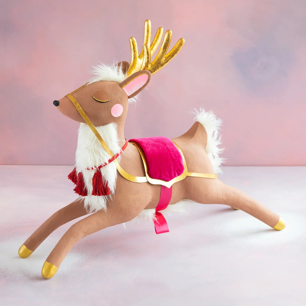 Glitterville Papa’s Reindeerfeaturing a hand-painted tan reindeer with tall metallic gold antlers, closed eyes, rosy pink cheek, fluffy white mane and tail, and gold-painted hooves. The reindeer wears a bright pink velvet saddle trimmed in gold, a red scarf with tassels, and a delicate gold harness. Shown leaping across a soft pink and white snowy background.