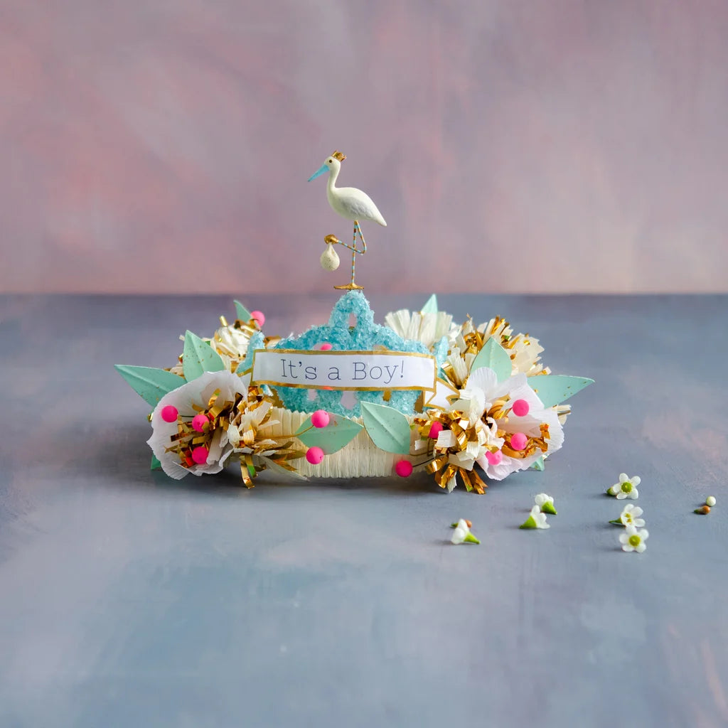 Glitterville Paper Mache Blue Royal Stork Tiara featuring a blue and white floral design, gold accents, and a small crowned stork topper with an “It’s a Boy!” sign.