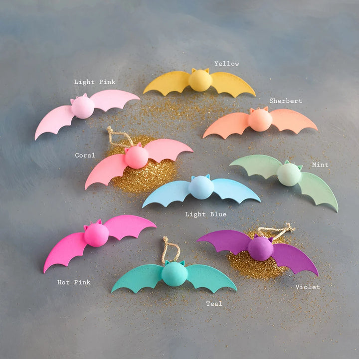 Rainbow Bat Glitterville pastel Rainbow Bat ornament set featuring whimsical bat decorations in pink, hot pink, yellow, peach, mint, blue, teal, and purple with coordinating round accents and gold glitter details. Each ornament is labeled by color for easy distinguishing. 