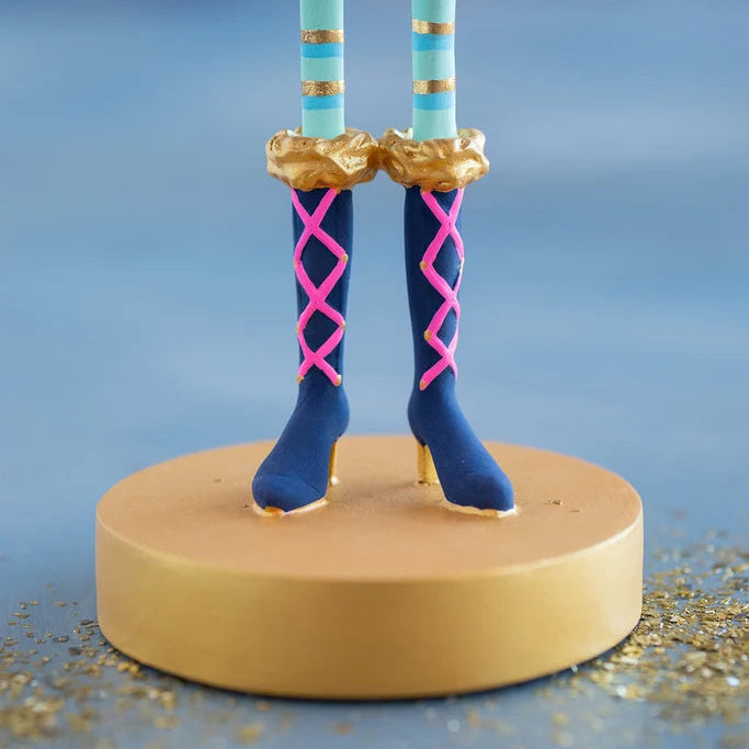 Close-up of a Glitterville Pinky Itchy Witchy tabletop doll showing turquoise legs with metallic gold stripes, navy blue knee-high witch boots featuring bright pink crisscross lacing and gold accents, and ruffled metallic gold boot cuffs. The doll stands on a round gold display base surrounded by scattered gold glitter against a soft blue studio backdrop.