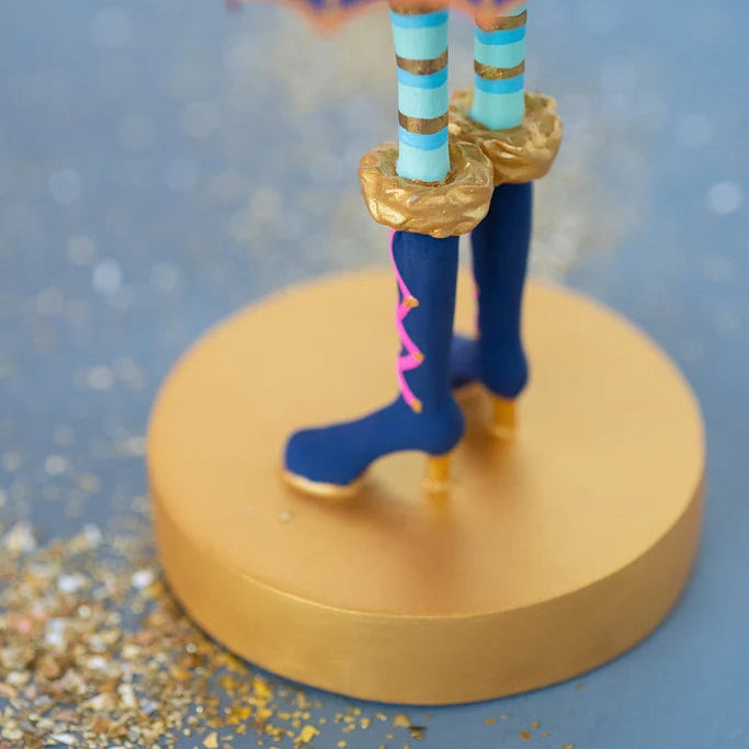 Close-up of a Glitterville Pinky Itchy Witchy tabletop doll showing navy blue witch boots with bright pink crisscross lacing and metallic gold details, turquoise legs with gold stripes, and ruffled gold boot cuffs. The boots stand on a round gold display base surrounded by scattered gold glitter against a soft blue studio backdrop.