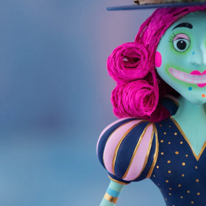 Close-up of a Glitterville Pinky Itchy Witchy tabletop doll featuring bright pink curled hair, a green face with painted eyes and a wide pink smile, and a navy blue dress with pink-and-gold striped puff sleeves and gold polka dots against a soft blue studio backdrop