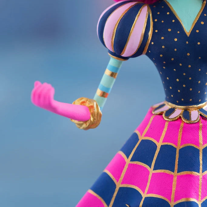 Close-up of a Glitterville Pinky Itchy Witchy tabletop doll showing the detailed navy blue and pink dress, gold polka dots, striped puff sleeve, gold-trimmed waist, pink and navy scalloped skirt with metallic gold outlines, and pink arm with a gold cuff against a soft blue studio backdrop
