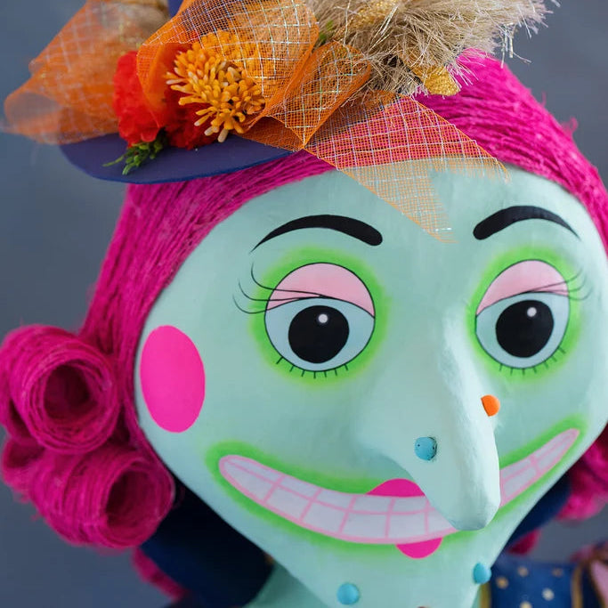Close-up of a Glitterville Pinky Itchy Witchy tabletop doll showing her mint green face, oversized black-and-white eyes with bright green and pink eye makeup, long pointed nose, pink cheeks, wide toothy smile, and vibrant hot pink curled hair. Her navy witch hat is decorated with metallic gold trim, fluffy tan accents, and an orange plaid mesh bow with colorful flowers.