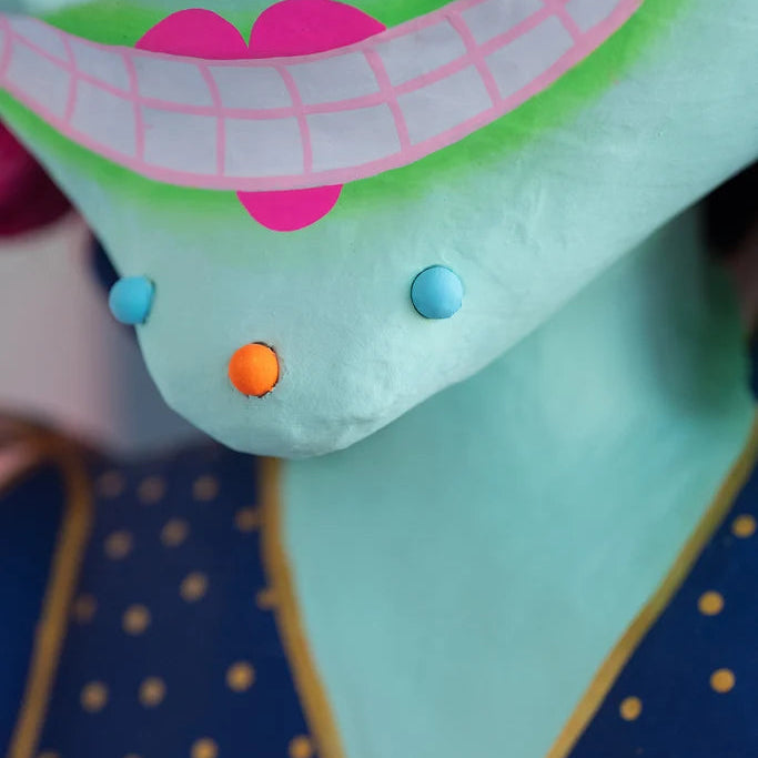 Close-up detail of Glitterville Pinky Itchy Witchy showing her mint green face, wide pink-lined toothy smile, bright pink lips, and small blue and orange decorative accents. The navy dress with gold polka dots is visible in the background.