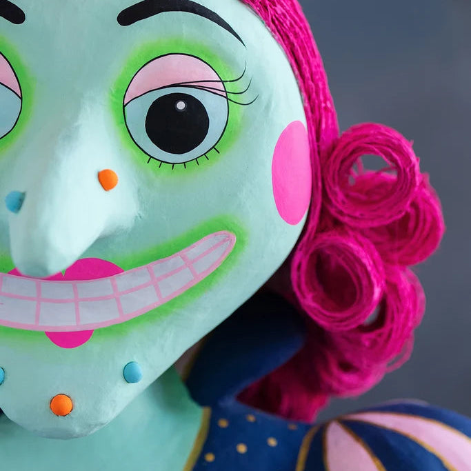 Close-up of a Glitterville Pinky Itchy Witchy tabletop doll featuring her mint green face, large expressive eyes with bright green and pink makeup, pointed nose, pink cheek, wide toothy smile, colorful face accents, and vibrant hot pink curled yarn hair. Part of her navy and gold polka-dot dress and pink-and-navy striped sleeve is visible against a soft blue-gray background.