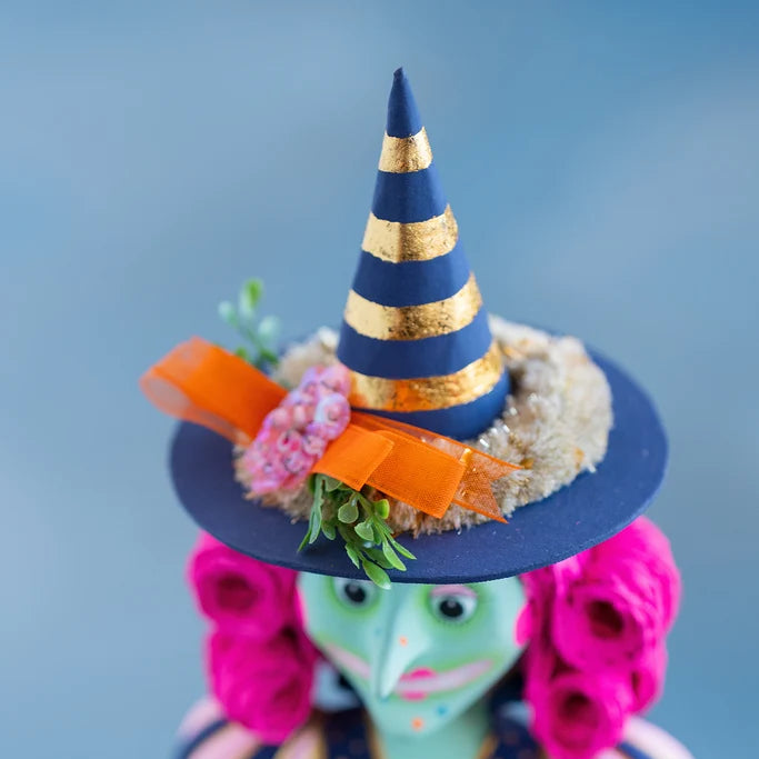 Close-up of Glitterville Pinky Itchy Witchy’s tall navy blue and metallic gold striped pointed witch hat, decorated with a wide orange ribbon, pink fabric flowers, cream textured trim, and green foliage, with her bright pink curled hair and green face softly visible below against a blue studio backdrop