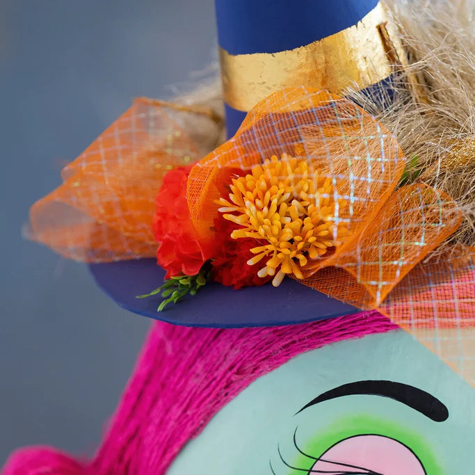 Close-up of a Glitterville Pinky Itchy Witchy tabletop doll featuring a navy blue pointed witch hat with metallic gold stripes, fluffy tan trim, and a decorative orange plaid ribbon bow with red and yellow flowers and greenery. Bright pink hair and part of the doll’s green face are visible beneath the hat against a soft blue studio backdrop.