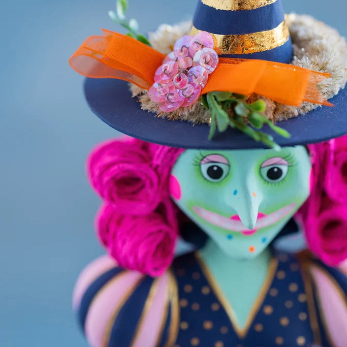 Close-up of Glitterville Pinky Itchy Witchy featuring a navy and gold striped pointed witch hat decorated with a bright orange ribbon, pink fabric flowers, dried floral accents, and green leaves, with her green face, pink curled hair, and navy polka-dot dress visible against a soft blue backdrop
