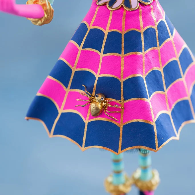 Close-up of a Glitterville Pinky Itchy Witchy tabletop doll showing the pink and navy blue spiderweb-patterned skirt with metallic gold outlines and a small gold spider embellishment at the center, with striped legs and gold-trimmed shoes visible against a soft blue studio backdrop