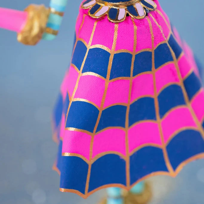 Close-up of a Glitterville Pinky Itchy Witchy tabletop doll featuring a bright pink and navy blue spiderweb-patterned skirt with metallic gold web detailing, scalloped hem, and a gold-trimmed waistband with alternating pink and navy accents. The doll’s striped turquoise arm and gold cuff are visible against a soft blue studio backdrop.