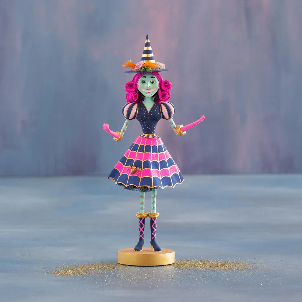 Glitterville Pinky Itchy Witchy tabletop witch figure featuring a green face, bright pink curled hair, pointed navy and gold striped witch hat, navy dress with pink and gold web-patterned skirt, striped stockings, pink-laced navy boots, and gold accents, displayed on a round gold pedestal against a soft blue and pink studio backdrop