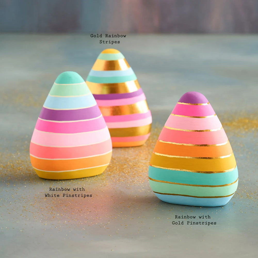 Glitterville rainbow-striped candy corn decorations shown in three color variations: rainbow with white pinstripes, gold rainbow stripes, and rainbow with gold pinstripes. The pastel and neon candy corn feature pink, coral, yellow, mint, aqua, and purple tones on a softly textured tabletop with scattered gold glitter.