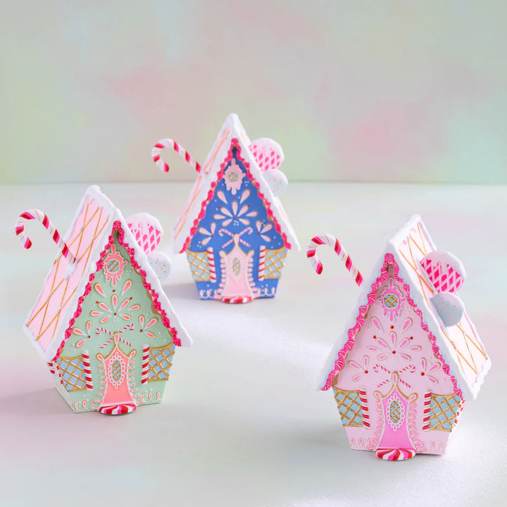 Glitterville rainbow gingerbread cottage tabletop Christmas display featuring three colorful pastel gingerbread houses in pink, blue, and green, each decorated with intricate white, pink, and gold icing, candy-cane accents, gold lattice details, peppermint trim, decorative windows, and snowy white icing. A whimsical pastel holiday scene perfect for Christmas decorating and collectible seasonal displays.