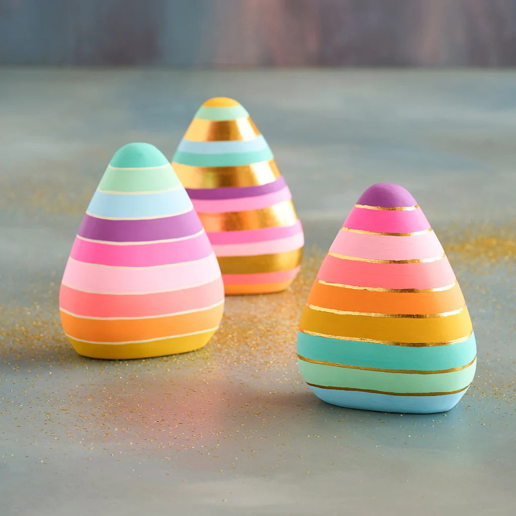 Glitterville set of three pastel and neon rainbow-striped candy corn decorations, featuring pink, coral, yellow, mint, aqua, and purple stripes accented with metallic gold bands. The three pieces are arranged on a softly textured tabletop with colorful glitter scattered around them against a muted blue and pink background.