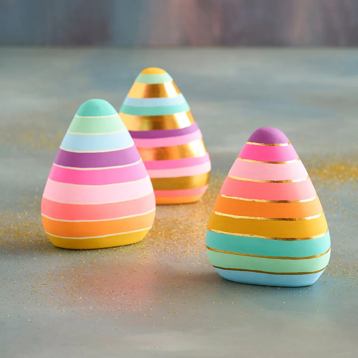 Glitterville set of three pastel and neon rainbow-striped candy corn decorations, featuring pink, coral, yellow, mint, aqua, and purple stripes accented with metallic gold bands. The three pieces are arranged on a softly textured tabletop with colorful glitter scattered around them against a muted blue and pink background.