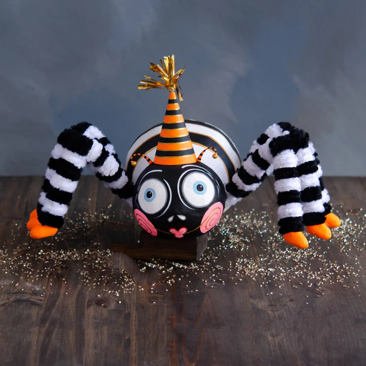 Glitterville Spinderella the Spider large display Halloween decoration featuring a whimsical black spider with oversized blue-and-white eyes, pink spiral cheeks, a heart-shaped smile, fuzzy black-and-white striped legs, bright orange feet, and a black-and-orange striped party hat with metallic gold accents.