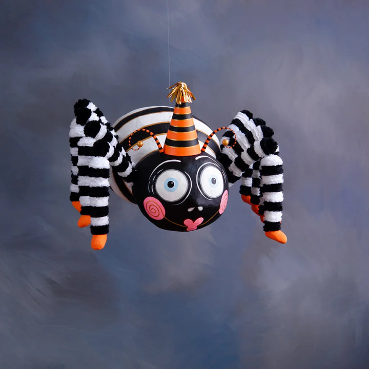 Glitterville Spinderella the Spider tabletop Halloween decoration featuring a whimsical black spider with oversized blue-and-white eyes, pink spiral cheeks, a heart-shaped smile, fuzzy black-and-white striped legs, orange feet, and a black-and-orange striped party hat with gold accents.