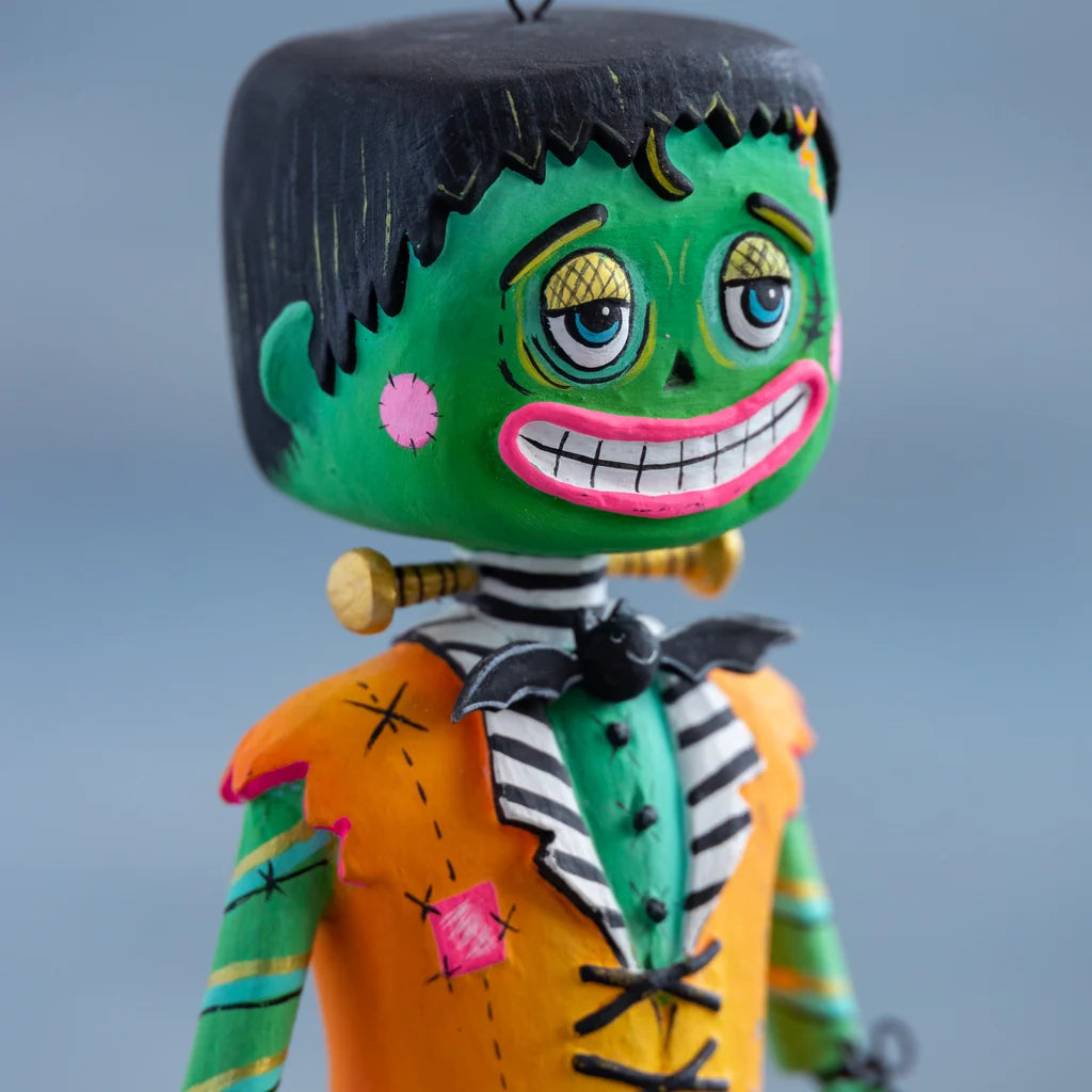 Close-up of a hand-painted Glitterville Swanky Franky Halloween ornament, showing the detailed green Frankenstein face, expressive eyes, stitched details, pink cheek, wide toothy grin, and orange jacket.