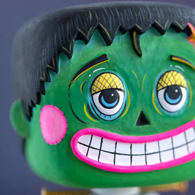 Close-up of a hand-painted Glitterville Swanky Franky Halloween ornament head, highlighting the detailed green Frankenstein face, blue eyes, crosshatched eyelids, stitched cheek, black hair, and bright pink toothy grin.