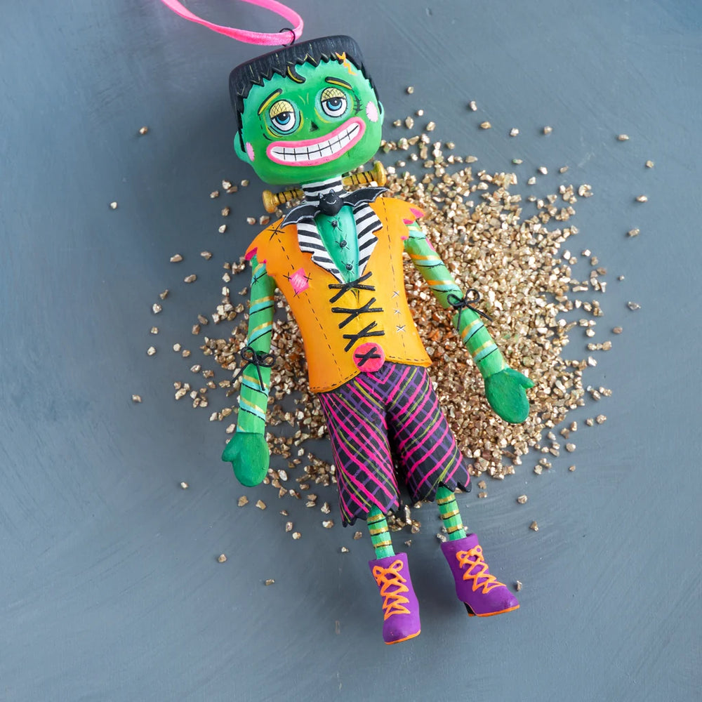 Glitterville Swanky Franky Halloween ornament lying on a bed of sparkling gold glitter, featuring a colorful green Frankenstein character in an orange vest, striped pants, and purple boots.
