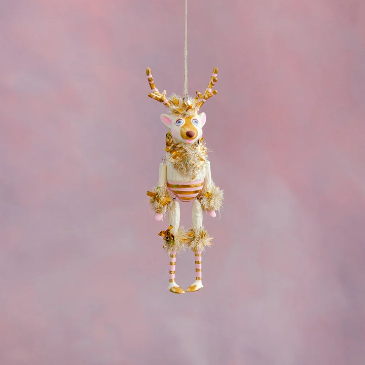 Gold and blush La Renne reindeer Christmas ornament with gold antlers, pink accents, striped body, and gold tinsel details hanging against a soft pink holiday backdrop.