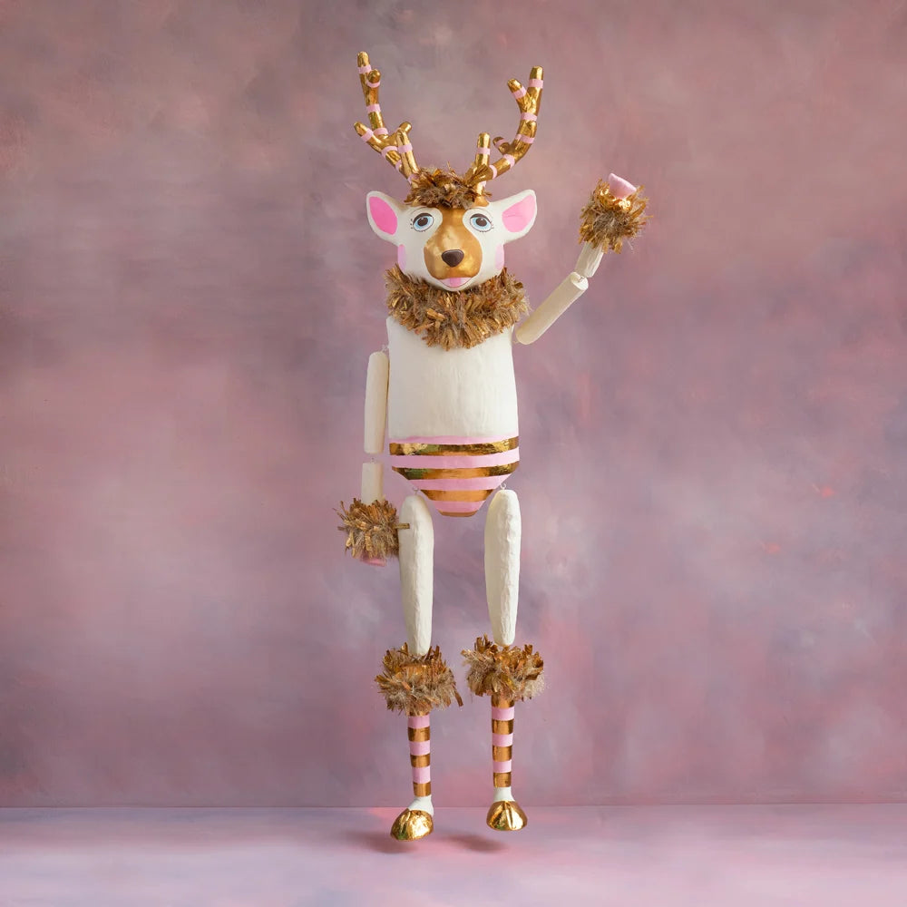 Gold and blush La Renne reindeer tabletop display with white body, pink accents, striped gold antlers, gold tinsel trim, and raised arm against a soft pink holiday backdrop.