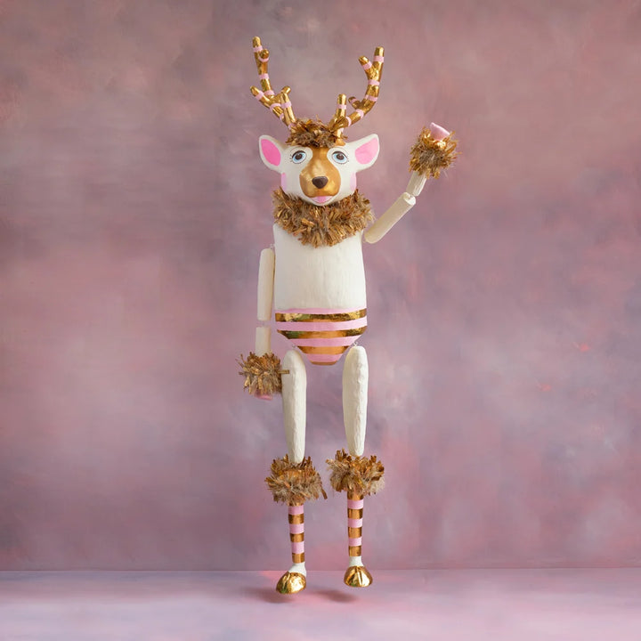 Gold and blush La Renne reindeer tabletop display with white body, pink accents, striped gold antlers, gold tinsel trim, and raised arm against a soft pink holiday backdrop.