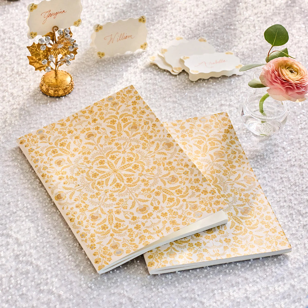 Gold floral journal styled as a wedding guest book with elegant place cards, floral decor, and gold accents on a sparkling tablescape.