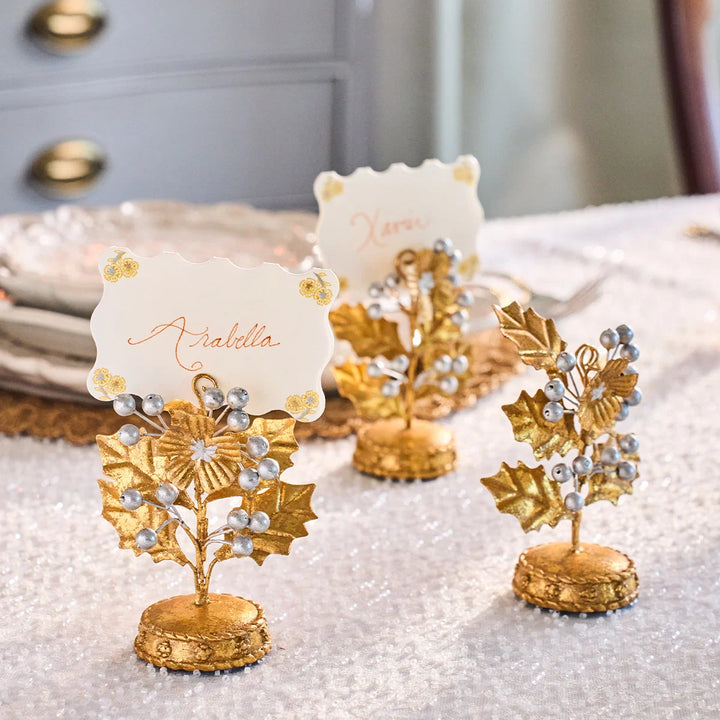Gold holly and silver berry place card holders displaying name cards on an elegant white and gold holiday table setting.