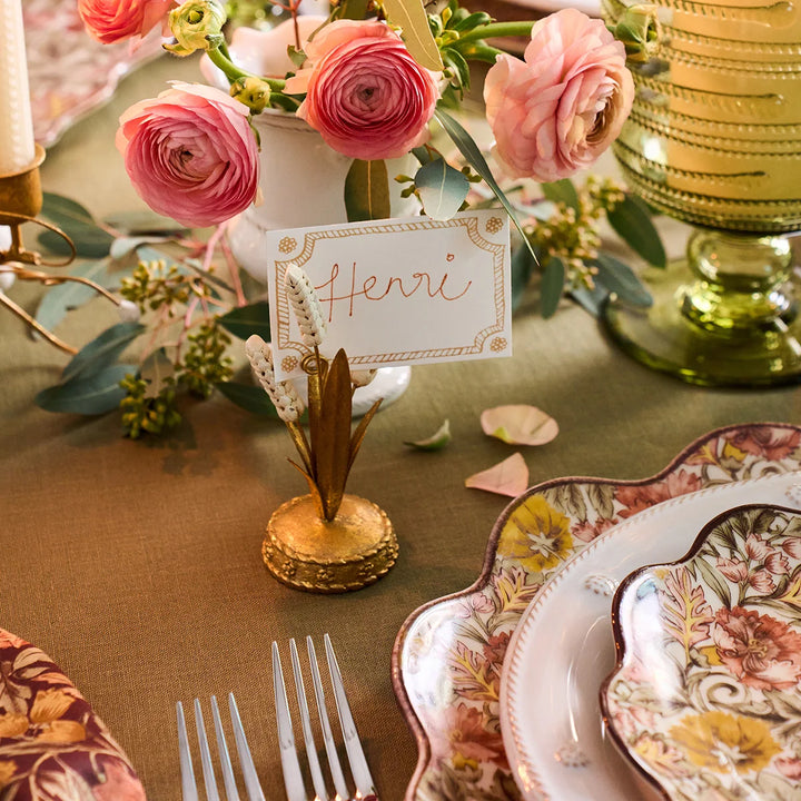 Gold Wheat Place Card Holder displaying a handwritten name card beside pink flowers and floral dinnerware on an elegant tablescape