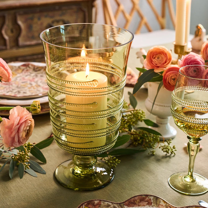 Green glass hurricane candle holder with glowing pillar candle styled among pink flowers and elegant tabletop decor.