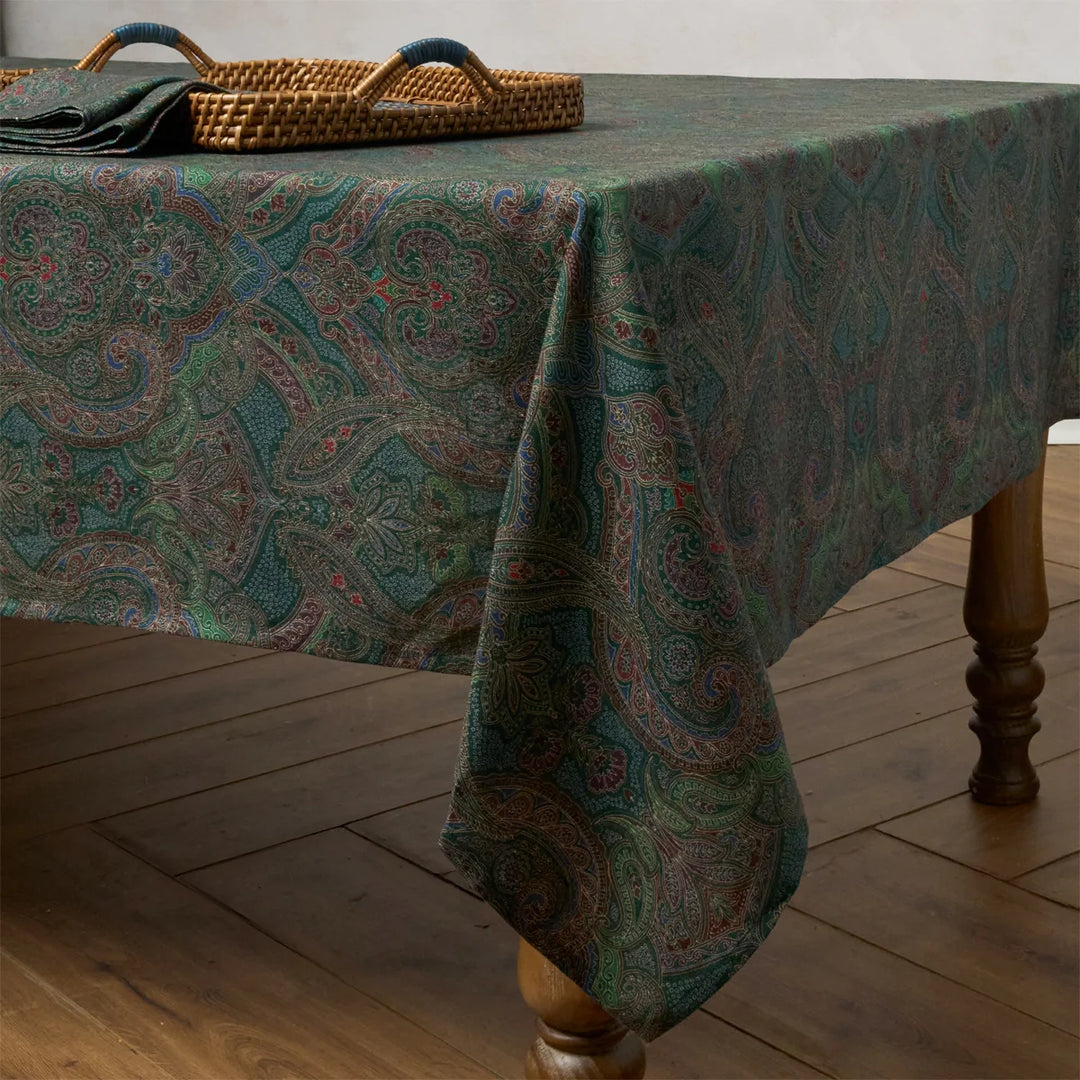 Green paisley tablecloth draped over a wood dining table, showcasing an elegant jewel-tone paisley and floral pattern.