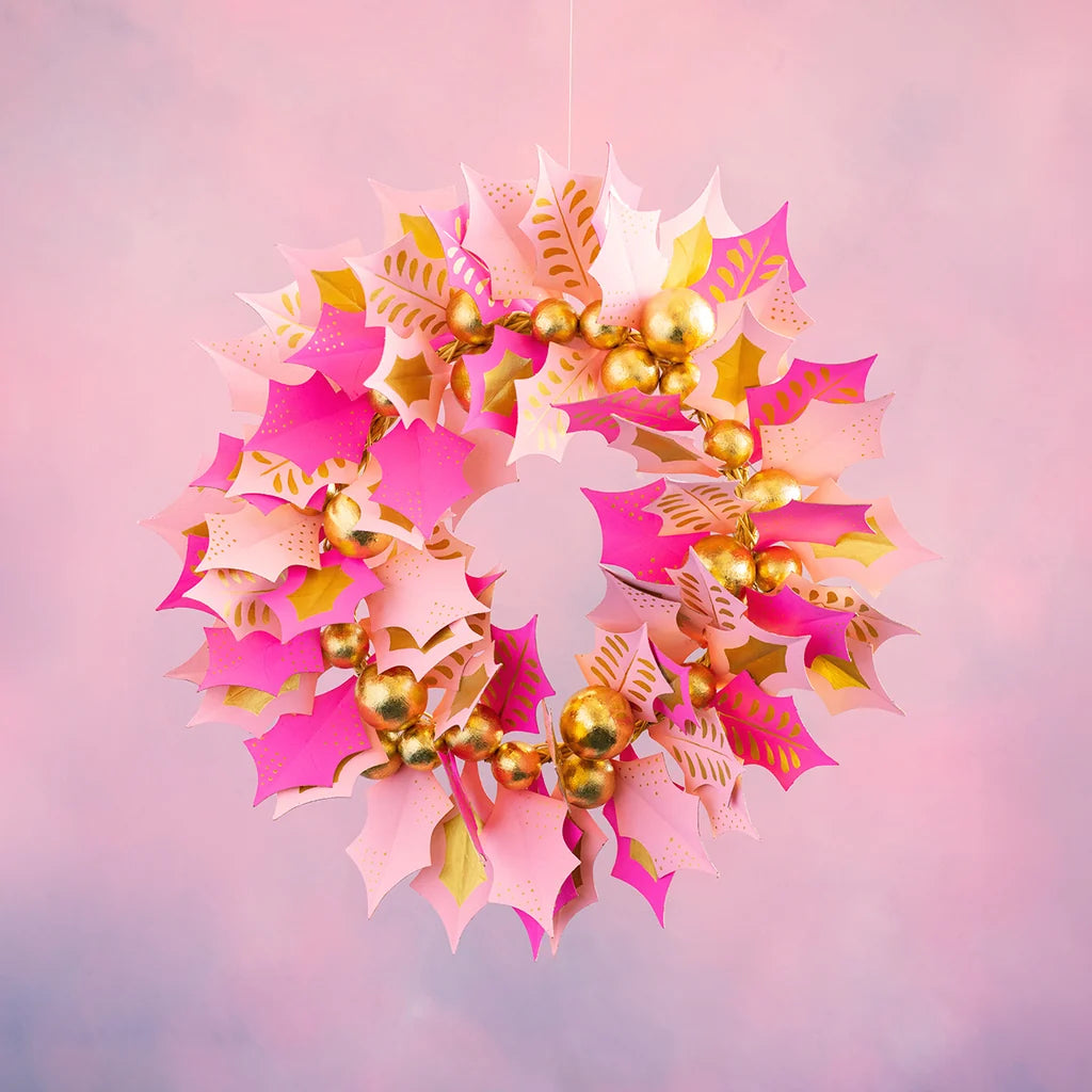 Pretty pink and gold holly wreath with layered prickly holly leaves, gold berries, and pink accents against a soft pink backdrop.