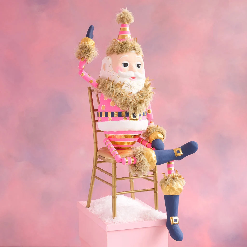 Hot pink and gold Papa Noel Christmas display seated on a wooden chair, wearing a pink striped suit with navy blue boots and gold accents, against a soft pink holiday backdrop.