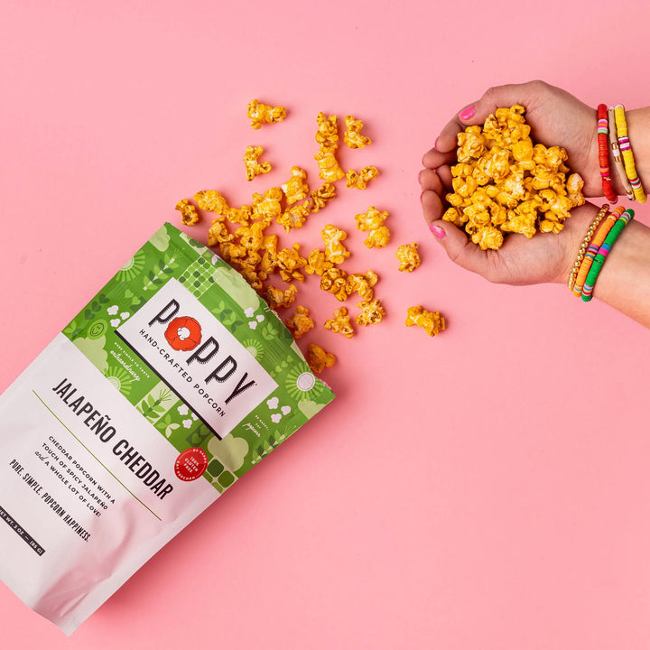 Poppy Jalapeño Cheddar Popcorn with seasoned popcorn held in hands against a pink background.