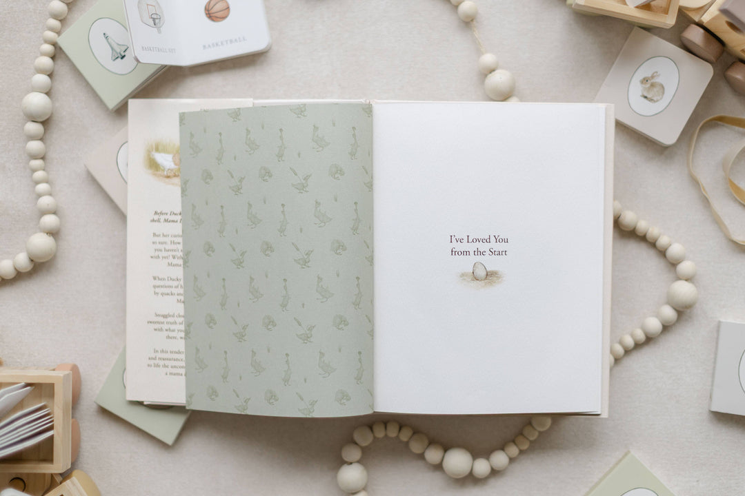 An open baby keepsake book laid flat on a neutral fabric surface, surrounded by wooden beads, small cards, and baby accessories. The left page has a pale sage green botanical pattern, and the right page is white with the printed text &