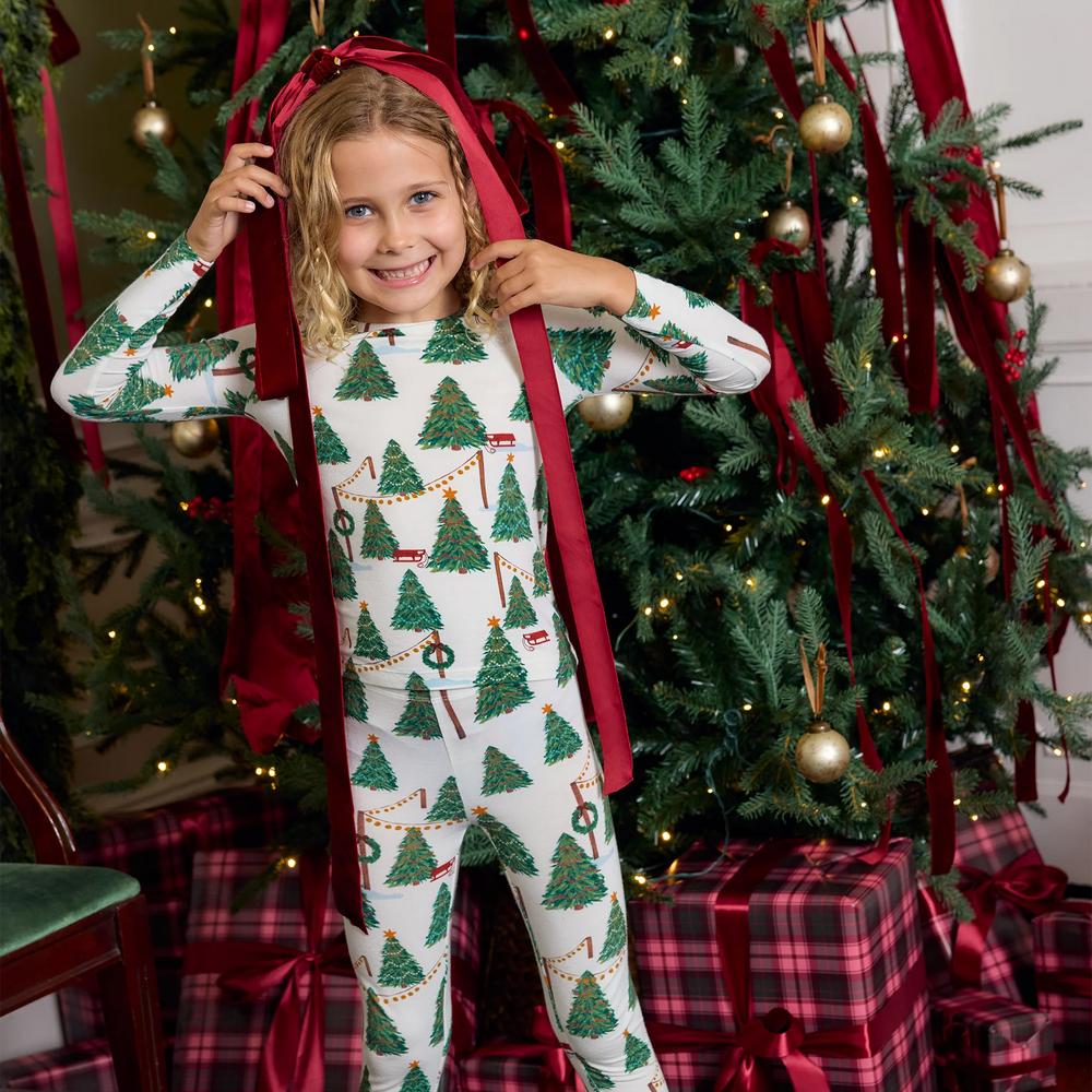 Girl wearing Christmas Tree Farm bamboo pajamas while smiling beside a decorated Christmas tree with red velvet ribbons.