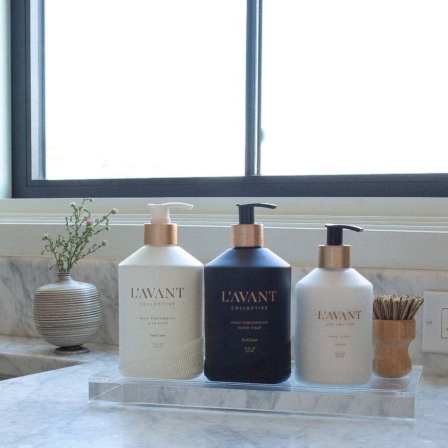 Large clear lucite organizer tray holding dish soap, hand soap, hand lotion, and a scrub brush on a marble countertop.