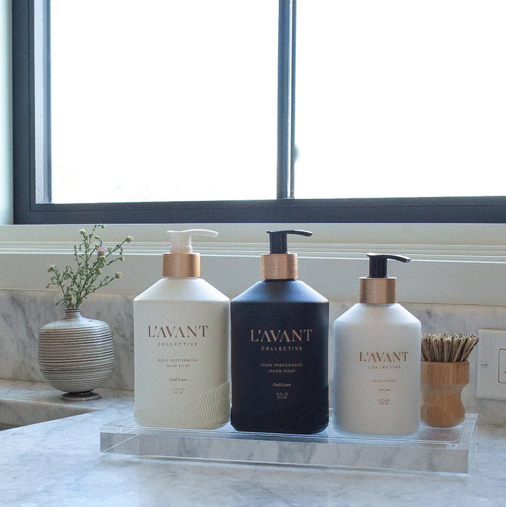 Large clear lucite organizer tray holding dish soap, hand soap, hand lotion, and a scrub brush on a marble countertop.