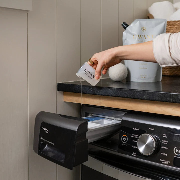 Silicone laundry detergent measuring cup being used to pour liquid detergent into a washing machine detergent dispenser.