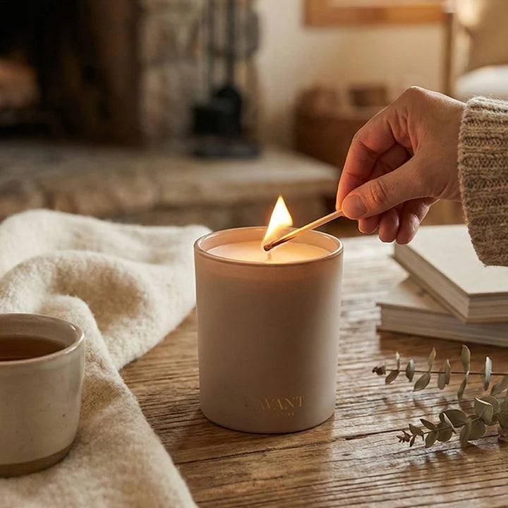 L’AVANT Collective Ambre Santal scented candle being lit with a match in a cozy home setting.