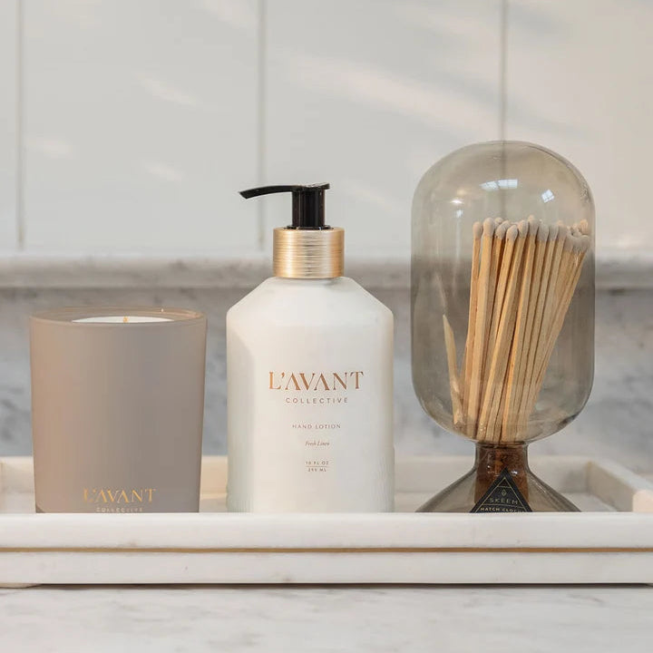 L’AVANT Collective Ambre Santal candle styled with hand lotion and decorative matches on a marble vanity tray.
