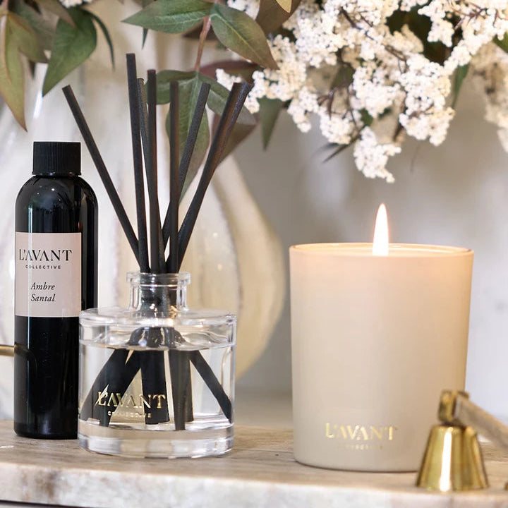 L’AVANT Collective Ambre Santal candle displayed with reed diffuser and fragrance refill beside white flowers.