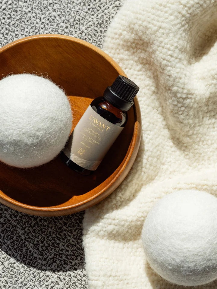L’AVANT Collective Ambre Santal Laundry Fragrance Oil displayed with a white wool dryer ball in a wooden bowl.